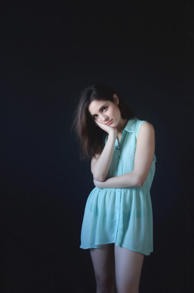 Mariana Esquivel a model with a contemplative expression stands against a dark backdrop wearing a light blue sleeveless dress with a ruffled collar.